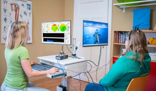Neurofeedback-Sitzung in der Praxis: Patientin mit EEG-Sensor vor dem Feedback-Monitor, Therapeutin am Auswertungsbildschirm