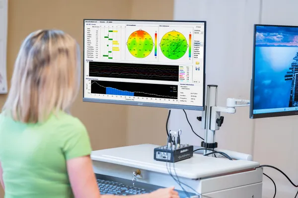 Monitor mit Neurofeedback-Software: EEG-Hirnkarten, Frequenzbänder und das NeuroMaster-Gerät auf dem Tisch